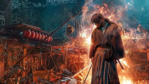 Kenshin stands with his sword in front of a burning traditional building at night
