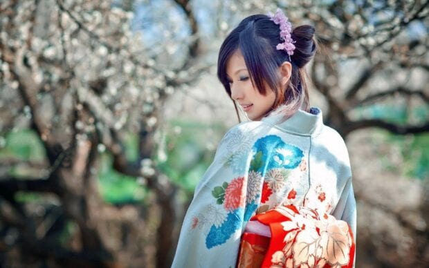 A woman wearing a colorful kimono with floral patterns stands outdoors among trees blooming in spring