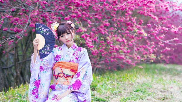 A woman wearing a kimono holding a decorative fan in a garden with blooming pink flowers