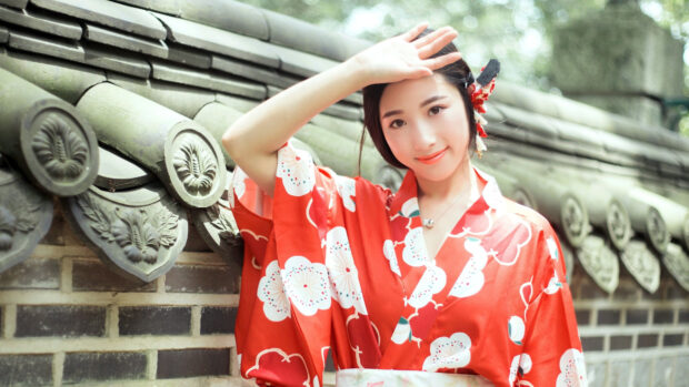 A young woman wearing a red kimono with floral patterns stands by a traditional stone wall covering her eyes from the sunlight
