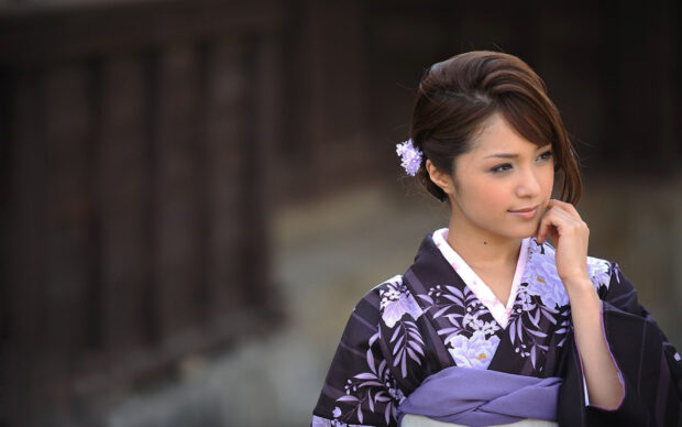 A woman wearing a purple kimono stands thoughtfully outdoors with a flower in her hair