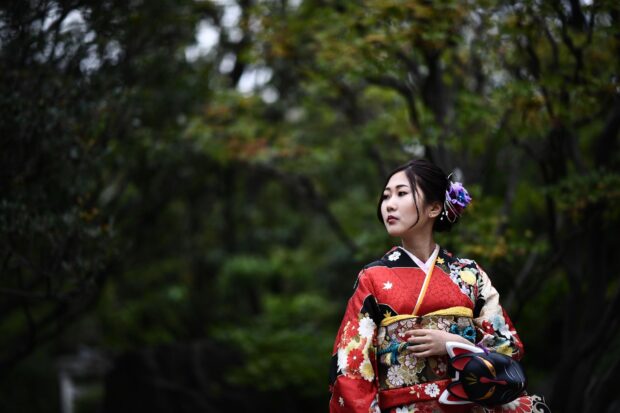 A woman wearing a traditional kimono stands in a lush garden holding a fox mask