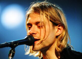 Kurt Cobain singing into a microphone on stage with his eyes closed and long blonde hair