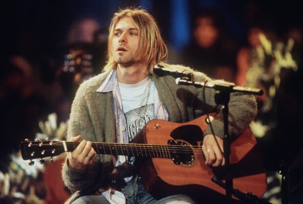 Kurt Cobain performing an acoustic guitar session wearing a cozy cardigan and a graphic t shirt