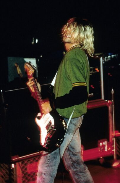 Kurt Cobain playing guitar on stage wearing a green shirt and jeans