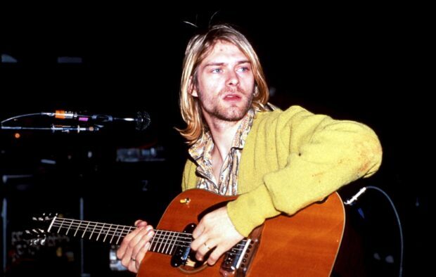 Kurt Cobain playing guitar with focused expression on stage in a yellow sweater