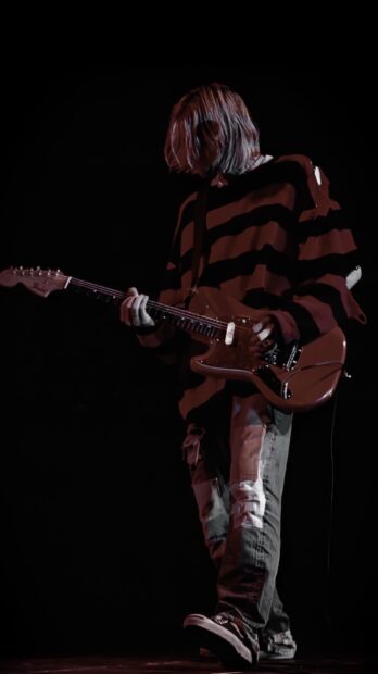 Kurt Cobain playing guitar on stage wearing a striped sweater and patched jeans