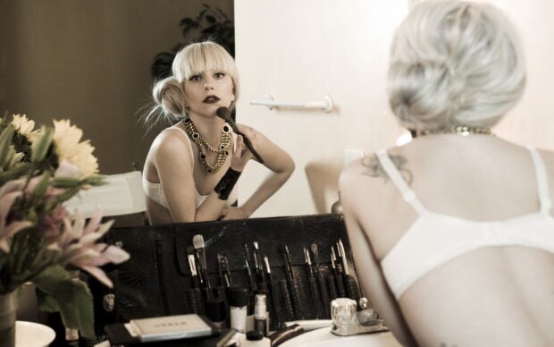 Lady Gaga applying makeup in front of a mirror with makeup brushes and flowers nearby