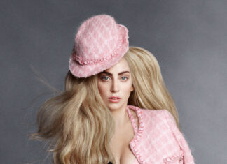 Lady Gaga wearing a pink hat and jacket with long blonde hair on a gray background