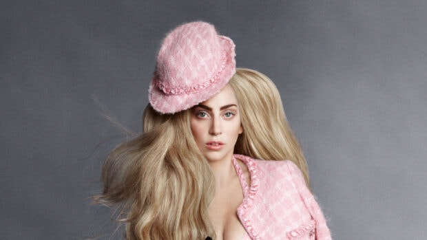 Lady Gaga wearing a pink hat and jacket with long blonde hair on a gray background