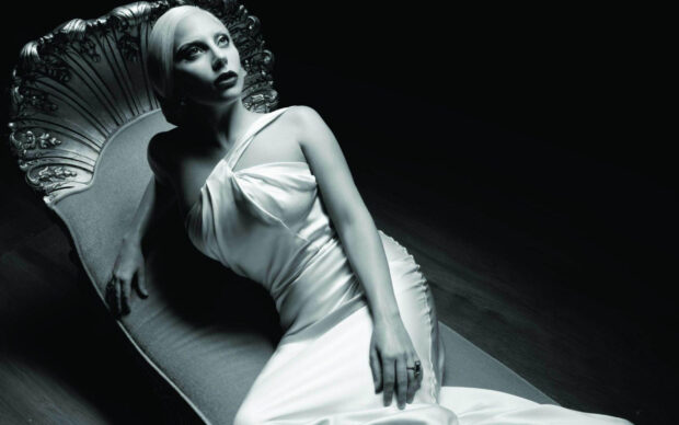 Lady Gaga wearing a white satin dress reclining on an ornate chair in a dark room
