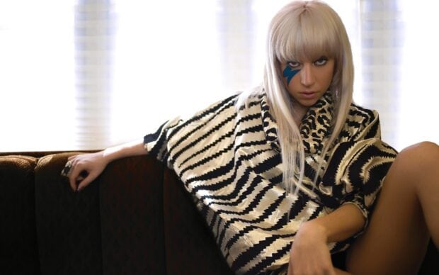 Lady Gaga wearing a patterned outfit with a blue lightning bolt on her face seated on a couch