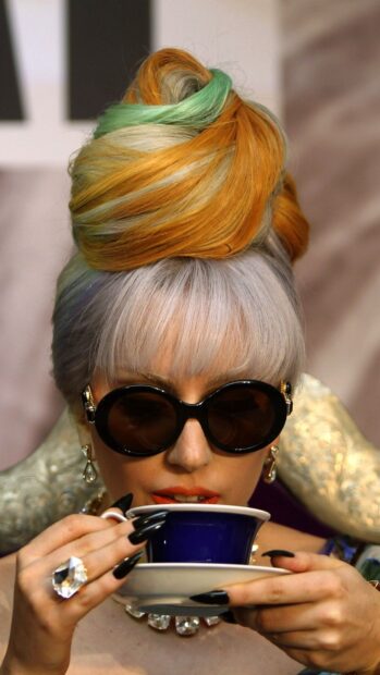 Lady Gaga with colorful hair and black sunglasses holding a blue cup close to her lips