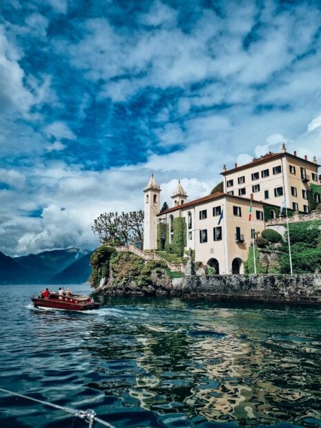 A boat sailing near a historic building on Lake Como with mountains and cloudy sky
