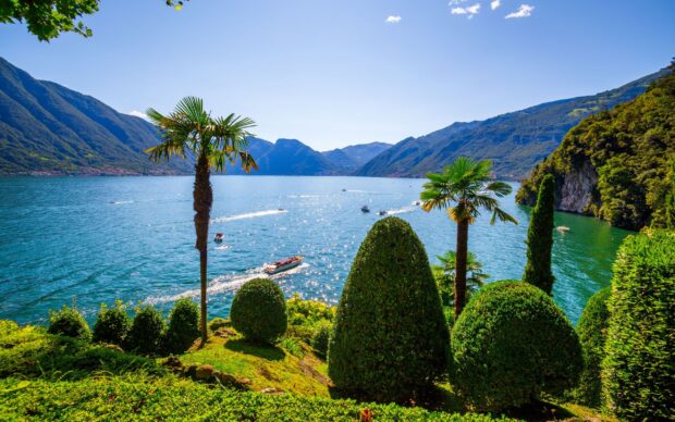 A scenic view of Lake Como with green manicured bushes and boats on the sparkling blue water surrounded by mountains