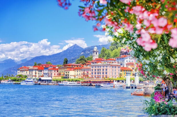 Colorful buildings along the Lake Como shore with flowers in the foreground and mountains in the background