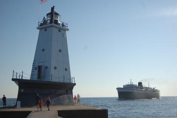 People walking near a lighthouse on Lake Michigan with a ship approaching in the background