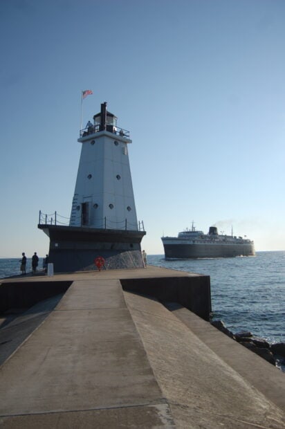 A lighthouse on a pier with people fishing and a ship sailing on Lake Michigan in the background