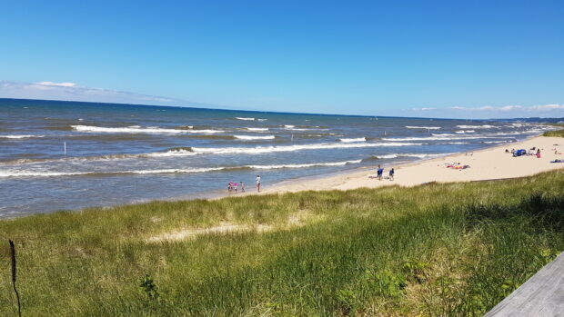A scenic view of Lake Michigan with grassy dunes and people enjoying the sandy beach on a sunny day