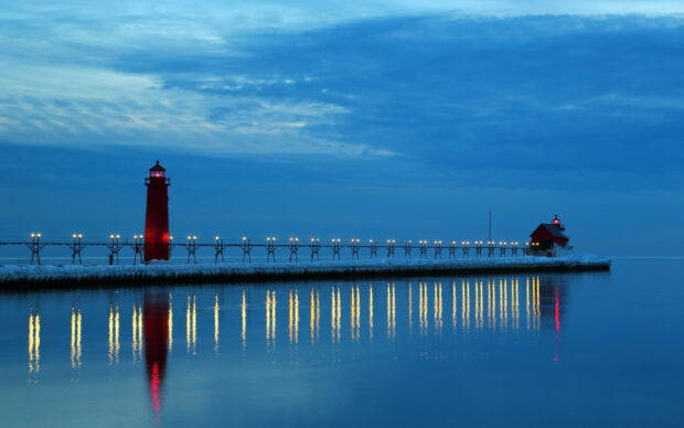 Red lighthouse and pier along Lake Michigan reflecting lights on calm water at dusk