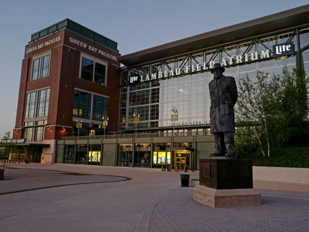 A statue stands in front of Lambeau Field with Green Bay Packers signage visible on the building
