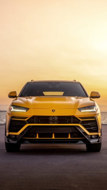 A yellow Lamborghini Urus is parked facing forward at sunset with a calm ocean background