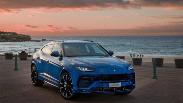 Blue Lamborghini Urus parked near the beach during sunset with surfers in the background