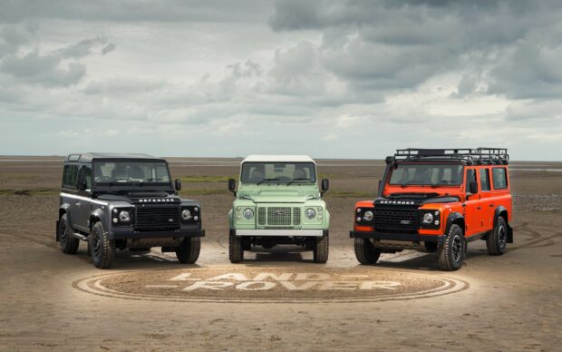 Three Land Rover Defender vehicles in black green and orange parked on a sandy beach with Land Rover logo in the sand