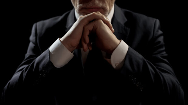 A lawyer with a beard wearing a suit and white cuffs with hands clasped thoughtfully