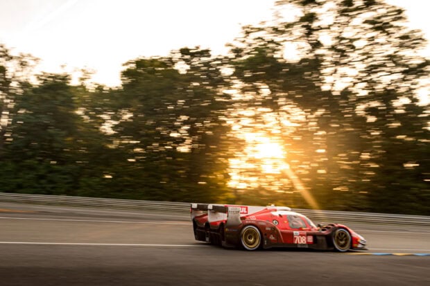 A red Le Mans race car driving fast on the track during sunset with trees in the background