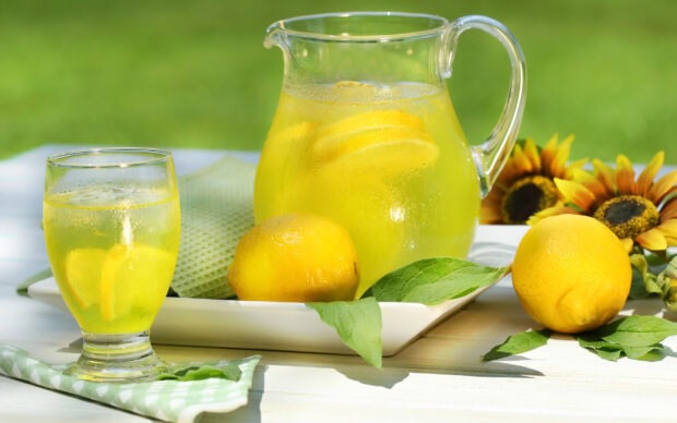 A pitcher and glass of fresh lemonade with lemons and green leaves on a white table outdoors