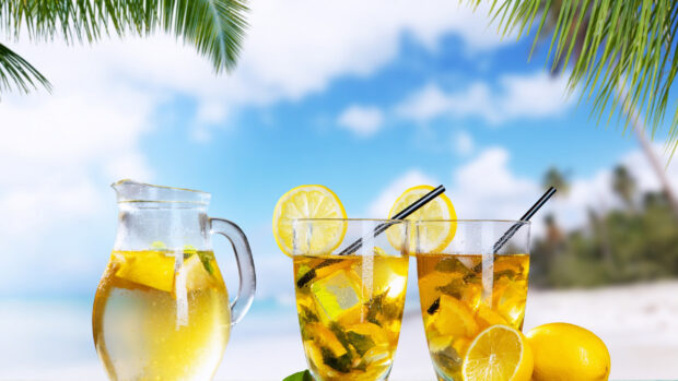 A pitcher and two glasses of lemonade with lemon slices on a tropical beach background
