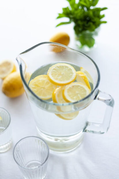Fresh lemon slices floating in a clear pitcher of water on a white table with blurred lemons and greens in the background