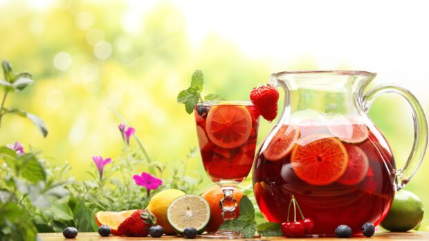 A refreshing lemonade drink with fruit slices and fresh mint leaves on a wooden table surrounded by berries and flowers
