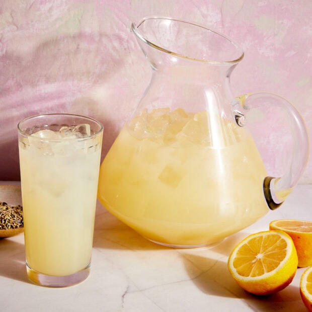 A glass of lemonade with ice cubes next to a pitcher of lemonade and fresh lemon halves on a marble surface