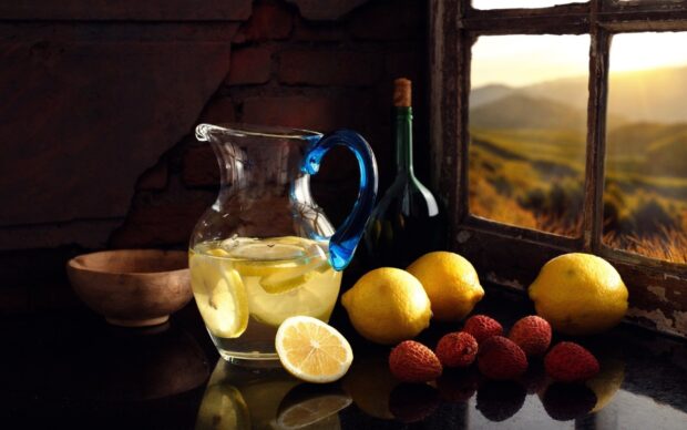 A glass pitcher filled with lemonade and lemons placed beside lychee fruits on a dark surface near a rustic window