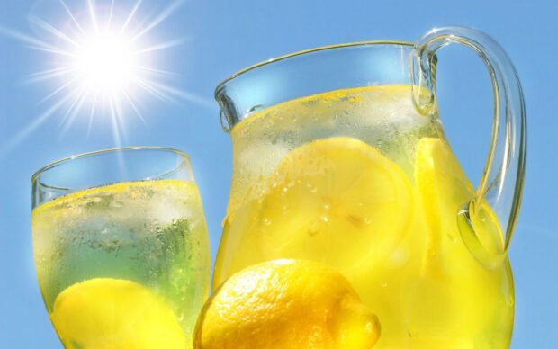 A glass and pitcher of lemonade with fresh lemon slices under bright sunlight