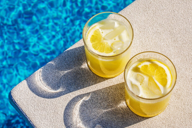 Two glasses of iced lemonade with lemon slices by a poolside surface