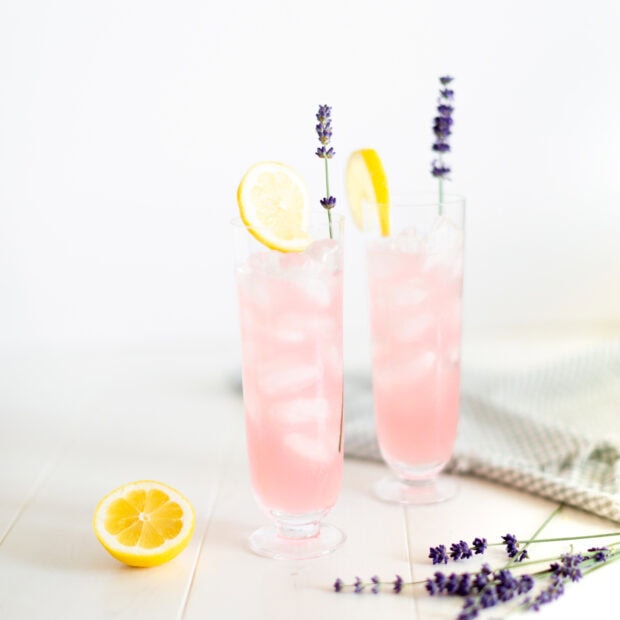 Two refreshing lemonade drinks with lemon slices and lavender on a white table with a lemon half and lavender flowers nearby