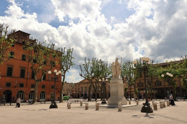 A statue surrounded by trees and historic buildings in Lucca Italy under a bright cloudy sky