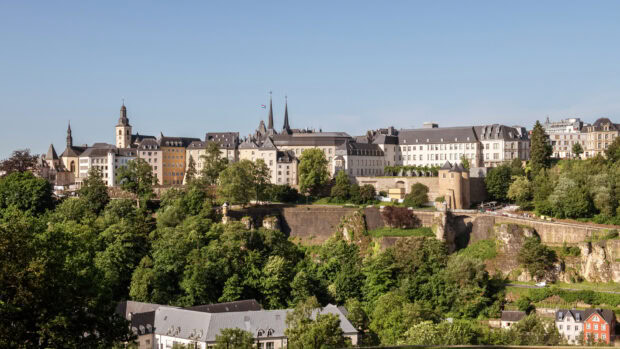 A scenic view of Luxembourg city with historic buildings and lush green trees on a clear sunny day