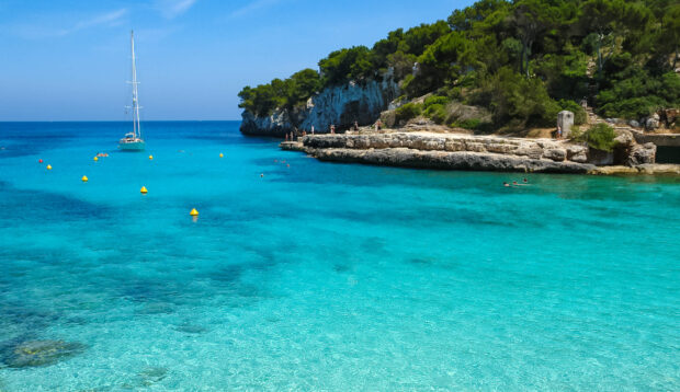 Crystal clear turquoise water near rocky coastline in Mallorca with sailing boat and swimmers