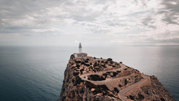 Winding road leading to a historic lighthouse on the Mallorca coastline with cloudy sky