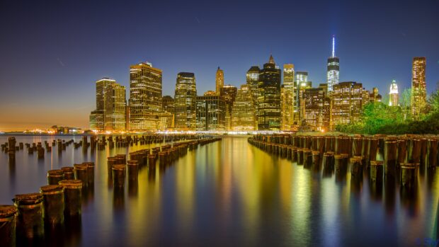 Night view of Manhattan skyline with old wooden posts in water reflecting city lights