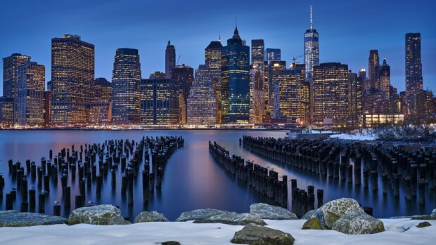 Snowy waterfront with wooden pillars in the water and Manhattan cityscape at dusk