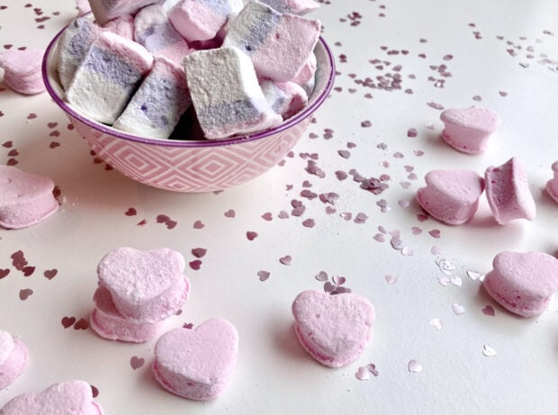 A bowl filled with pastel marshmallow treats surrounded by pink heart shaped marshmallows and confetti
