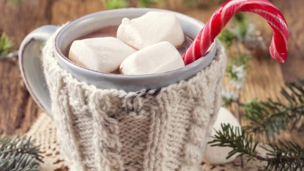 Hot chocolate with marshmallow and candy cane in a cozy knitted cover on a wooden table