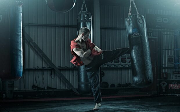 Male martial artist practicing high kick training in gym with punching bags
