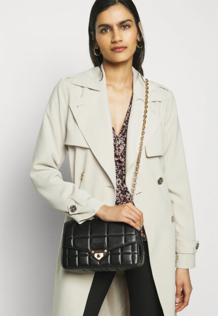 A woman wearing a beige coat holding a Michael Kors handbag with a gold chain strap