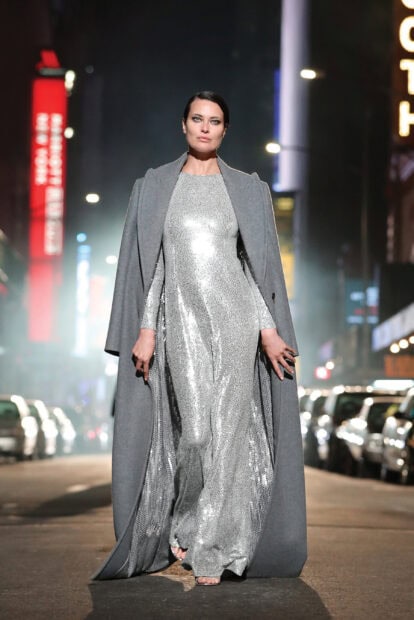 A model wearing a shimmering silver dress and grey coat on a city street at night with Michael Kors design